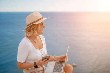 Fototapeta premium Freelance women sea working on the computer. Good looking middle aged woman typing on a laptop keyboard outdoors with a beautiful sea view. The concept of remote work.