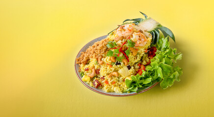 Freshly prepared pineapple fried rice served inside of half a pineapple like boat on a plate on a yellow background. Thai pineapple stuffed with rice, shrimp, chicken and vegetables on the table