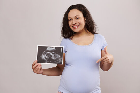 Happy pregnant expectant mother holding scan of her child in womb, smiling at camera and pointing finger at her belly, experiencing positive moments of her pregnancy and maternity, isolated background