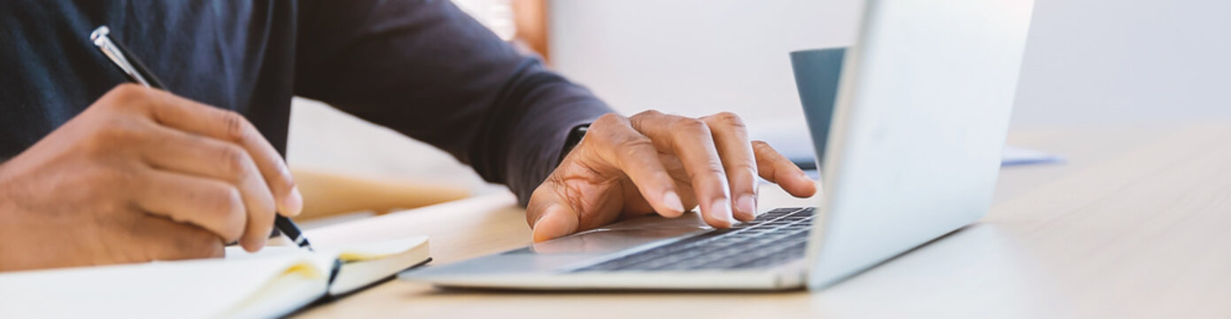 Closeup businessman hand holding pen writing on notebook, work on laptop computer in modern office. Student lecturing while studying online internet course. Write on journal diary banner (blur)