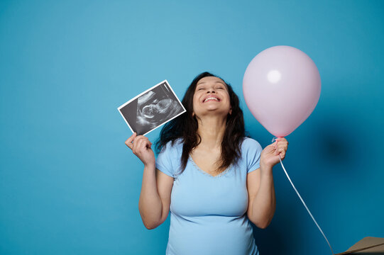 Overjoyed excited amazed pregnant woman, expectant mom expressing happiness and positive emotions, expecting baby girl, posing with pink balloon and ultrasound scan image over blue isolated background