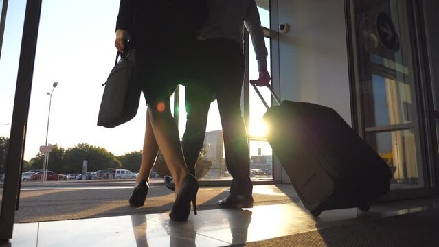 Business people leaving airport through automatic glass door with their luggage. Young man and woman in heels walking from terminal and roll suitcase on wheels. Concept of work trip. Slow motion