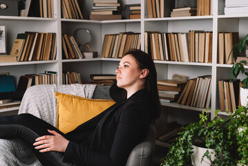 Happy dreamy young woman sitting in armchair and looking away. Concept of wellbeing and self-care.