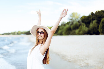 Fototapeta na wymiar Happy smiling woman in free happiness bliss on ocean beach standing with a hat, sunglasses, and rasing hands. Portrait of a multicultural female model in white summer dress enjoying nature during trav