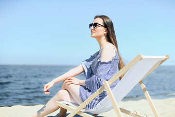 Happy young brunette woman relaxing on a wooden deck chair at the ocean beach while smiling and wearing fashion sunglasses. The enjoying vacation concept.