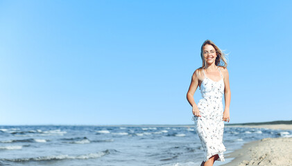 Fototapeta na wymiar Happy blonde woman in free happiness bliss on ocean beach standing straight. Portrait of a female model in white summer dress enjoying nature during travel holidays vacation outdoors.