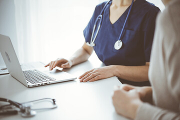 Doctor and patient sitting at the desk in clinic office. The focus is on unknown female physician's hands using a laptop computer, close up. Medicine concept.