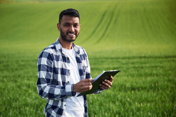 Standing and holding notepad. Handsome Indian man is on the agricultural field