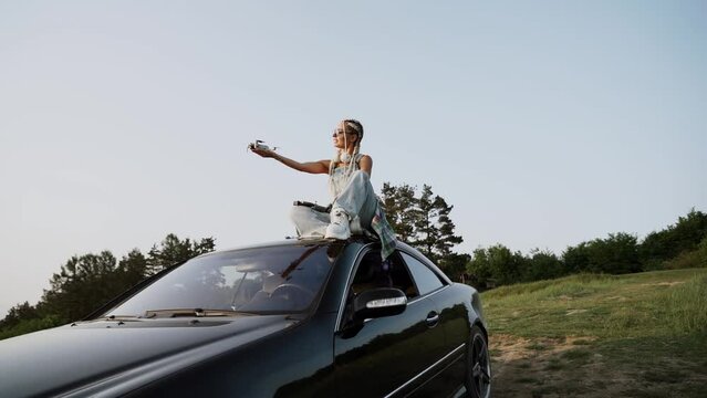 Stylish woman in glasses launches a drone at sunset sitting on the roof of a car. Drone pilot concept