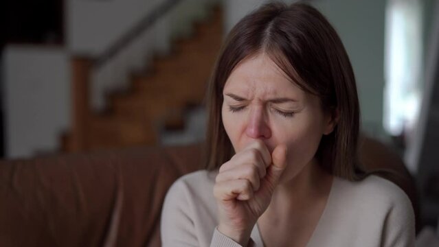 Close-up sick young woman with cold and flu coughing sitting in living room at home. Ill sad Caucasian lady looking away rubbing painful neck thinking