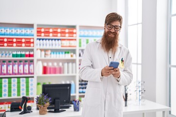 Young redhead man pharmacist using smartphone working at pharmacy