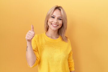 Young caucasian woman wearing yellow sweater doing happy thumbs up gesture with hand. approving expression looking at the camera showing success.