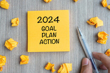 2024 Goal, Plan, Action word on yellow note with Businessman holding pen and crumbled paper on wooden table background. New Year New Start, Resolutions, Strategy  concept