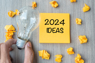 2024 Idea words on yellow note and crumbled paper with Businessman holding lightbulb on wooden...