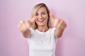 Young blonde woman standing over pink background approving doing positive gesture with hand, thumbs up smiling and happy for success. winner gesture.