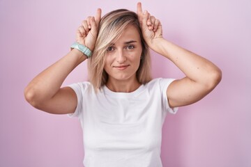 Young blonde woman standing over pink background doing funny gesture with finger over head as bull horns