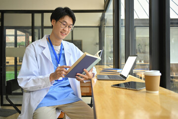 Smiling male medical student in glasses and white coat reading book and using laptop in hospital canteen