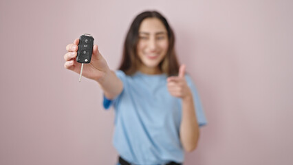 Young beautiful hispanic woman doing thumbs up pointing to key of new car over isolated pink background