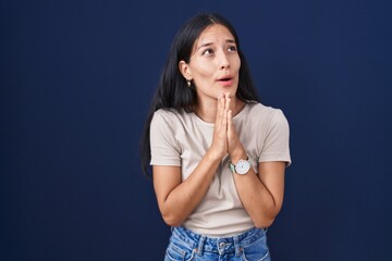 Young hispanic woman standing over blue background begging and praying with hands together with hope expression on face very emotional and worried. begging.