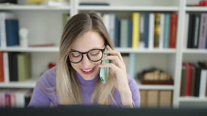 Young blonde woman student using computer talking on smartphone at library university