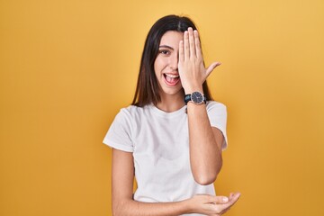 Young beautiful woman standing over yellow background covering one eye with hand, confident smile on face and surprise emotion.