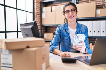 Young beautiful hispanic woman ecommerce business worker holding dollars using calculator at office