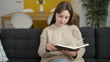 Young blonde woman reading book sitting on sofa at home