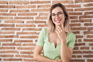 Young beautiful woman standing over bricks wall looking confident at the camera smiling with crossed arms and hand raised on chin. thinking positive.