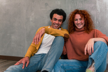 Multiracial friends sit on the floor, leaning against the wall, dressed in casual clothing, smiling