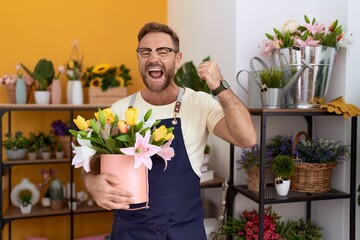 Middle age man with beard working at florist shop holding plant screaming proud, celebrating victory and success very excited with raised arms