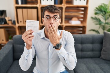Young hispanic man holding covid record card covering mouth with hand, shocked and afraid for...