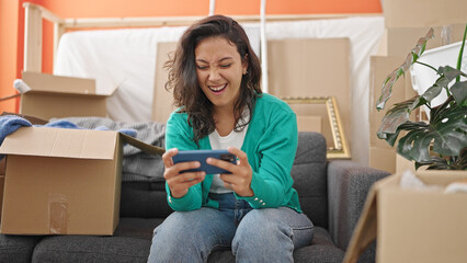 Young beautiful hispanic woman playing video game sitting on sofa at new home