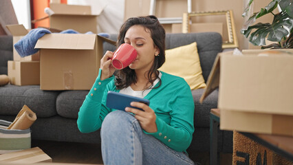 Young beautiful hispanic woman watching video on smartphone drinking coffee at new home