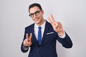 Young hispanic man wearing suit and tie smiling looking to the camera showing fingers doing victory sign. number two.