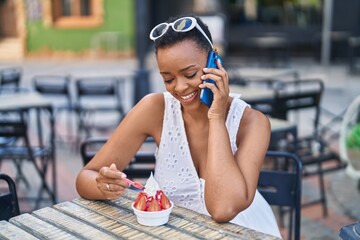 African american woman eating ice cream talking on smartphone at coffee shop terrace
