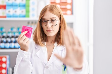 Young redhead woman working at pharmacy drugstore holding condom with open hand doing stop sign with serious and confident expression, defense gesture