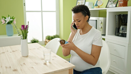 Young pregnant woman having medical phone consultation sitting on table at dinning room