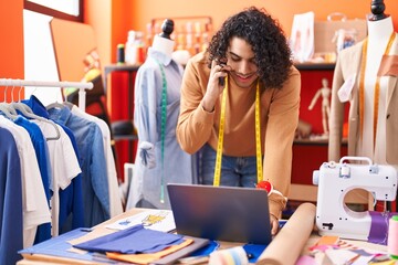 Young latin man tailor talking on smartphone using laptop at atelier