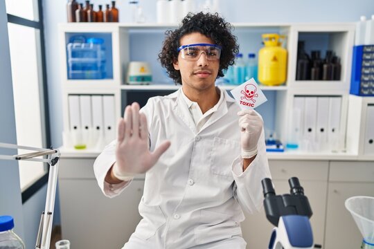 Hispanic man with curly hair working at scientist laboratory holding toxic banner with open hand doing stop sign with serious and confident expression, defense gesture