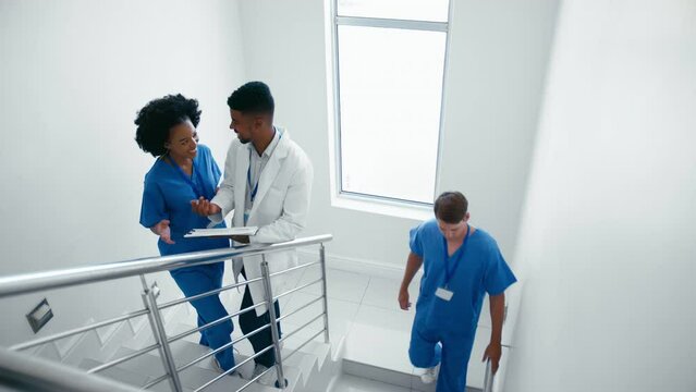 Male doctor wearing white coat discussing patient notes on clipboard with female nurse in scrubs on hospital stairs being passed by male colleague - shot in slow motion
