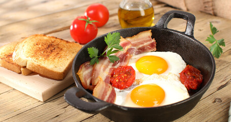 fried eggs with bacon and tomatoes in a frying pan. breakfast on the wooden table. sunny morning. country style. organic farm food