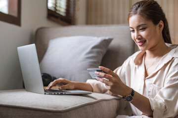 Young smiling Asian woman using smartphone buying online shopping by credit card while lying on sofa when relax in living room at home. Lifestyle latin and hispanic ethnicity women at house concept.