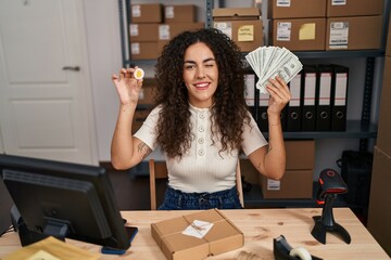 Young hispanic woman working at small business ecommerce holding money and bitcoin winking looking at the camera with sexy expression, cheerful and happy face.