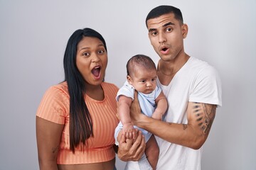 Young hispanic couple with baby standing together over isolated background in shock face, looking skeptical and sarcastic, surprised with open mouth