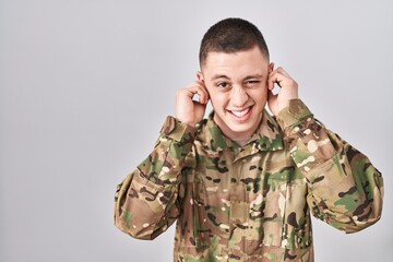 Young man wearing camouflage army uniform covering ears with fingers with annoyed expression for...