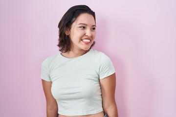 Hispanic young woman standing over pink background looking away to side with smile on face, natural expression. laughing confident.