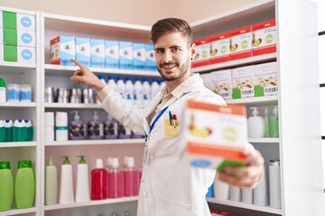 Young caucasian man pharmacist smiling confident holding vitamin package at pharmacy