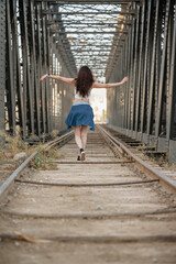 young woman with her back to the camera in casual clothes posing with her arms waving as she walks along old railway tracks over a bridge with large metal girders.