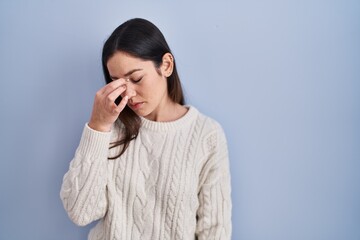 Young brunette woman standing over blue background tired rubbing nose and eyes feeling fatigue and...