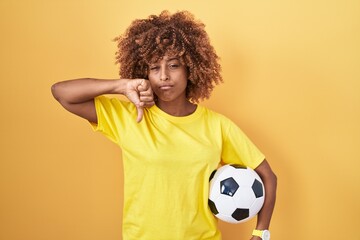 Young hispanic woman with curly hair holding football ball with angry face, negative sign showing...